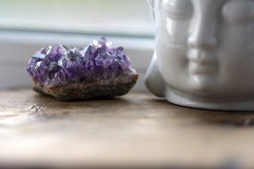 Purple and gemmy amethyst stone with White Buddha head on the windowsill background. Rough ametist...
