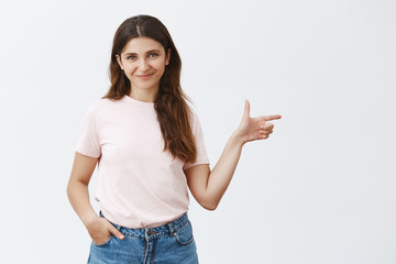 You would not regret seeing this. Portrait of charming cute and lovely brunette girl in pink t-shirt and jeans holding hand in pocket pointing right with finger gun and smiling friendly at camera