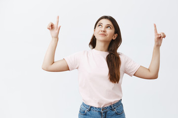 Indoor shot of cute and tender european woman wishing on star and dreaming raising index fingers pointing and looking up in sky with broad gentle smile having wish over gray background