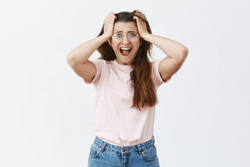 Studio shot of panicking desperate woman in despair wearing glasses and casual outfit screaming pulling hair out of head and staring shook and unhappy at camera having troubles and problems