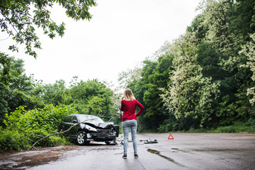 A young woman with smartphone by the damaged car after a car accident, making a phone call.