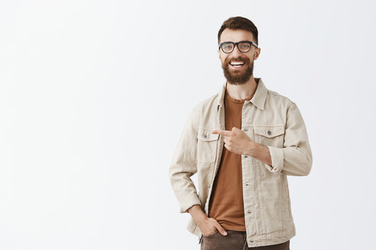 Studio Shot Of Friendly Joyful Handsome Guy With Long Beard And Stylish Moustache In Trendy Glasses And Outdoor Beige Jacket Laughing Out Loud Having Funny And Interesting Conversation Pointing Left