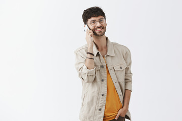 Making arrangements via smartphone. Friendly-looking successful and pleased male entrepreneur in glasses and trendy urban outfit holding hand in pocket and phone near ear grinning at camera