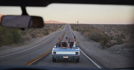 Group Of Friends On Road Trip Driving Classic Convertible Car Viewed Through Windshield Of...