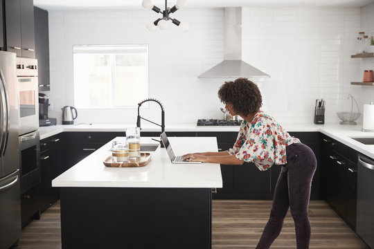 Woman At Home Standing At Kitchen Island Whilst Using Laptop