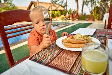 Cute boy picky eater frustrating over the dish with sausages, vegetables, spaghetti served with fresh pineapple juice on the table by the swimming pool in luxury hotel