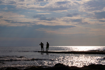 Due amici in cima a un molo in controluce, cielo nuvoloso, riflessi del solo sull'acqua del mare