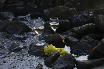 Weißwein und Weintrauben auf Steinen im fließenden Wasser bei Morgensonne.
