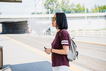 Vaping. Young beautiful woman smoking e-cigarette with smoke outdoors. Vapor concept.