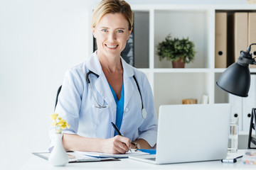 adult female doctor with stethoscope over neck writing in clipboard at table with laptop in office