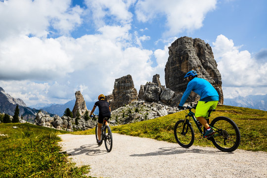 Tourist cycling in Cortina d'Ampezzo, stunning Cinque Torri and Tofana in background. Woman and man riding MTB trail. South Tyrol province of Italy, Dolomites.