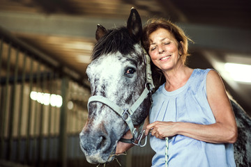 A senior woman standing close to a horse in a stable, holding it.
