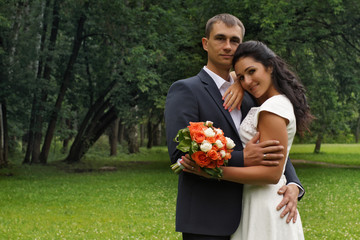 A pair of beautiful newlyweds stand on a glade in the middle of the park on a summer day