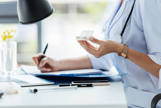 cropped image of female doctor writing in clipboard and holding pills at table in office