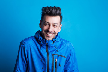 Portrait of a young laughing man in a studio with anorak on a blue background. © Halfpoint
