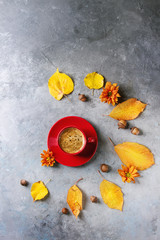 Red cup of black coffee with foam decorated by yellow autumn leaves, aster flowers and acorns over grey texture background. Flat lay, space. Seasonal background.