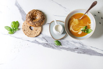 Bowl of vegetarian pumpkin carrot soup served with herbs, spoon, jug of cream, bagel bread, black...