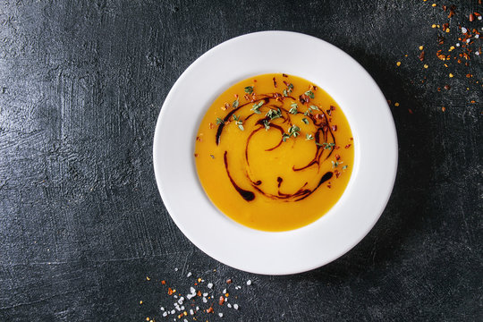 Plate of vegetarian pumpkin carrot soup decorated by balsamic vinegar and thyme served with chili pepper and salt over black texture background. Flat lay, space