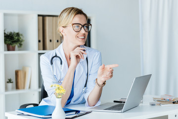 smiling female doctor in eyeglasses pointing by finger and talking at table with laptop in office