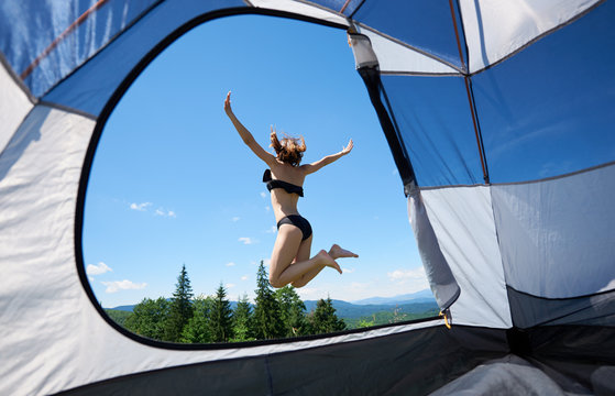 View from tent inside of sporty female jumping near the tent, enjoying summer day in the mountains. Camping lifestyle concept adventure summer vacations outdoor