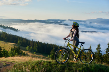 Fototapeta premium Sporty girl cyclist riding on yellow bicycle on a rural trail in the mountains, wearing helmet, enjoying valley view on sunny morning. Foggy mountains, forests on the blurred background