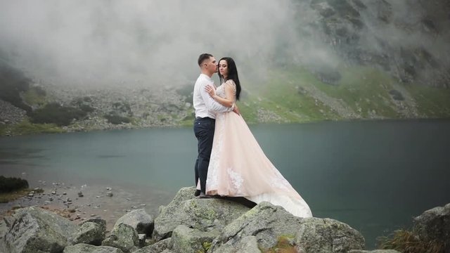 Lovely wedding couple stands near lake in mountains, hugging kissing on a cloudy day groom happy beautiful love smile beauty dress celebration party face bridge portrait summer young nature outdoor