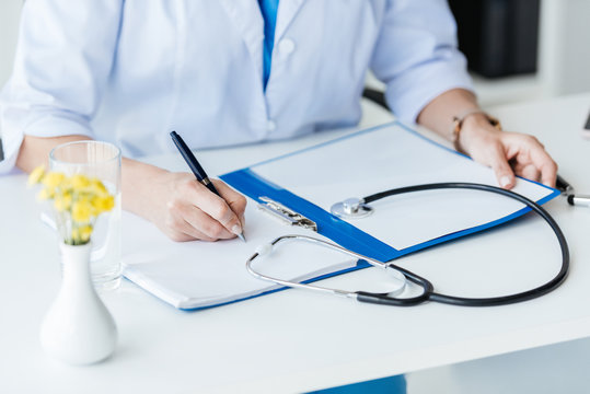 cropped image of female doctor writing in clipboard at table with stethoscope in office