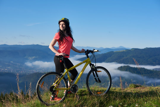 Attractive woman cyclist with yellow bike on a rural trail in the mountains, wearing helmet and red red t-shirt, enjoying morning haze in valley, forests on blurred background. Outdoor sport activity