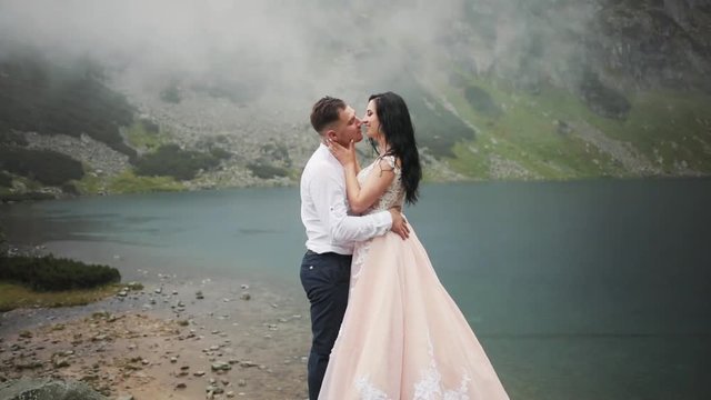 Portrait lovely wedding couple stands near lake in mountains, hugging kissing on a cloudy day groom happy beautiful dress celebration party face bridge summer young nature outdoor