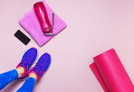 Young woman in sporting blue leggings, preparing for training. Bottle of water, yoga mat, phone on pastel pink background flat lay top view. Sports shoes, fitness, concept of healthy lifestile