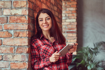 Smiling brunette girl student dressed in a plaid shirt and jeans holding digital tablet while leaning on a brick wall in a room with loft interior, looking at camera.