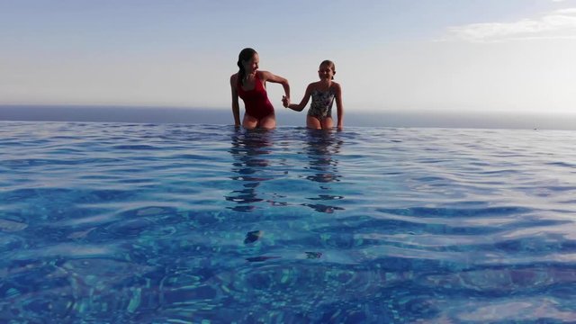 Two girls are jumping and swimming into infinity pool. 4K drone shot, seaview