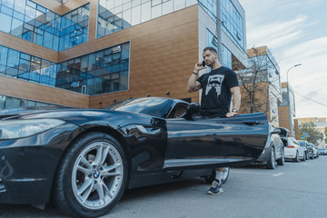 Young man getting out of the black car and talking by mobile phone. Side view.