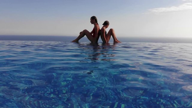 Two girls are sitting on the edge of an infinity pool. 4K drone shot, seaview