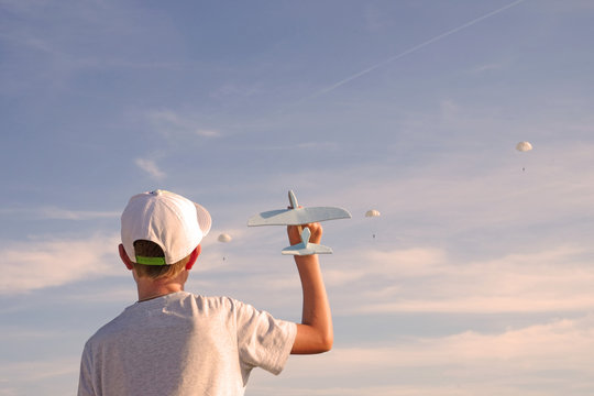 Back View Of Kid In Cap Holding Toy Plane And Standing Against Blue Sky With Skydivers