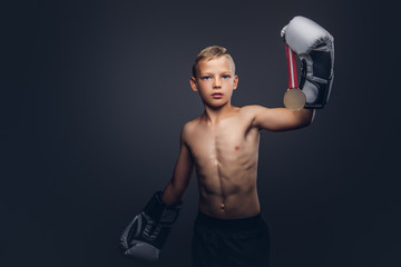 Young shirtless boy boxer in boxing gloves holds a golden medal posing in a studio.