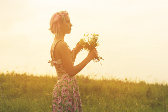 Young woman in dress with bouquet of flowers in hands at sunset in the field. Tinted warm image