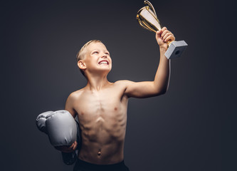 Joyful shirtless boy holds boxer gloves and the winner's cup.