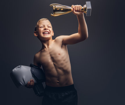 Joyful shirtless boy holds boxer gloves and the winner's cup.