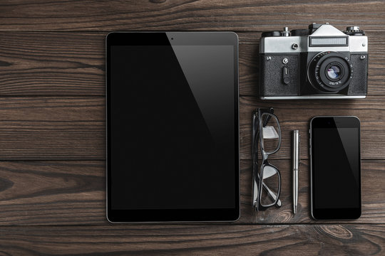 Old retro camera, tablet, silver pen, eyeglasses and smartphone on vintage wooden background. Top view, flat lay