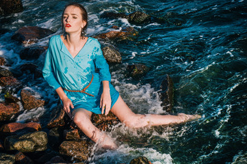 The fashion model sits on rocks and poses in the ocean water.