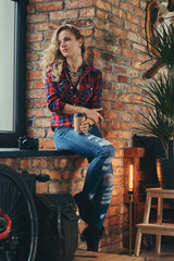 Obraz na płótnie Canvas Sensual blonde hipster girl with long curly hair dressed in a fleece shirt and jeans holds a cup of morning coffee sitting on a window sill at a studio with loft interior.