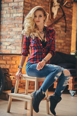 Fototapeta na wymiar Sensual blonde hipster girl with long curly hair dressed in a fleece shirt and jeans holds a cup of morning coffee sitting on a wooden stool at a studio with a loft interior.