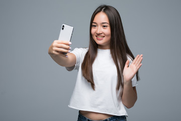 Naklejka premium Sociable beautiful asian girl taking selfie or speaking on video call using cell phone isolated over gray background