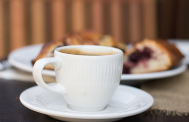 Close-up: white cup of coffee is on a wooden table. In the background, there are pieces of a delicious cherry pie.