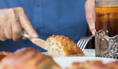 Savory delicious piece of baked pie is cut off and eaten by a man who sits at a table. He holds in his hands cutlery. A glass of tea is on a table.