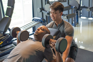 Trainer and man lying and lifting dumbbells in gym. Young black guy training. Bodybuilding concept.
