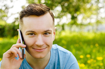 Young guy communicates on a mobile phone and sitting on green grass with dandelions on a background of trees