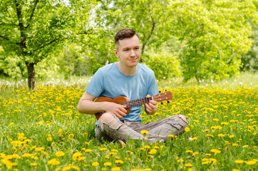 Naklejka na ściany i meble Young guy sits on a green grass with dandelions and plays on the ukulele and sings.