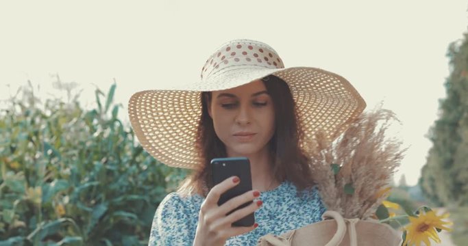 Woman takes selfie on cellphone at sunflower field. Sweet sexy girl in a blue dress and hat walks on a field of sunflowers , lifestyle 4K video shooting by handheld gimbal